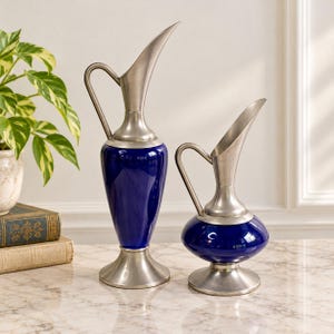 May include: Two decorative pitchers featuring deep blue glossy bodies and brushed silver-toned metal bases, handles, and spouts. The taller pitcher has a slender, elongated form, while the shorter pitcher has a rounded, bulbous base.