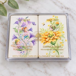 May include: A clear plastic case containing two decks of playing cards. The left deck features purple flowers and a fairy, while the right deck shows yellow flowers and a fairy. Text includes "TWO DECKS", "PLASTIC COATED PLAYING CARDS", "DEUX JEUX", "CARTES A JOUER", and "HOYLE PLAYING CARD CO. WILLOWDALE, ONT. M2H 3G2".