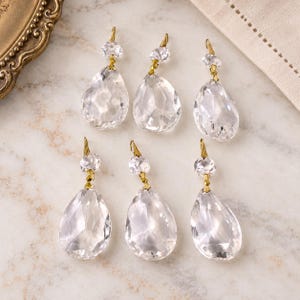 Peut inclure: Six pendentifs en forme de goutte en verre transparent à facettes, chacun surmonté d'une petite perle de cristal ronde. Les cristaux sont reliés par des épingles en métal doré, formant une structure d'ornement suspendue pour lustres.