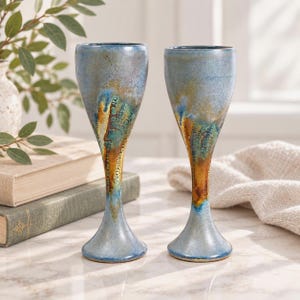 May include: Two tall ceramic goblets featuring a reactive glaze with a blend of light blue, deep teal, and warm amber tones. The surface displays a distinctive crackle texture that transitions from a smooth rim to a textured, earthy base.