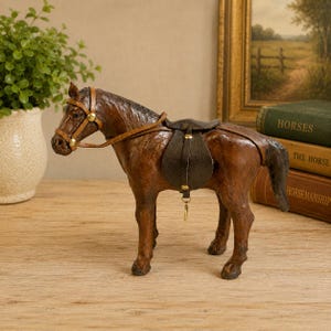 May include: A brown leather horse figurine featuring a dark brown saddle and a matching bridle with brass-colored hardware. The horse stands in a profile position, showcasing detailed stitching on the leather surface and a small metal ring attached to the saddle girth.
