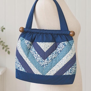 May include: A quilted fabric handbag featuring a chevron pattern of blue, white, and light blue floral and polka dot prints. The bag has a solid dark blue base, a gathered top opening, and two dark brown wooden beads at the ends of the handle.