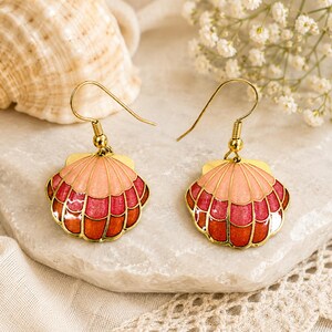 Peut inclure: Une paire de boucles d'oreilles en forme de coquillage avec des crochets dorés. Les boucles d'oreilles présentent un motif en éventail dans les tons de rose et de rouge, souligné d'or. Elles sont posées sur une surface blanche.
