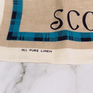 Puede incluir: Un pa&ntilde;o de cocina de lino color canela con un borde de cuadros azul oscuro y azul verdoso. El texto SCOT est&aacute; impreso en letras negras grandes y las palabras ALL PURE LINEN est&aacute;n impresas en letras negras peque&ntilde;as cerca del borde inferior.