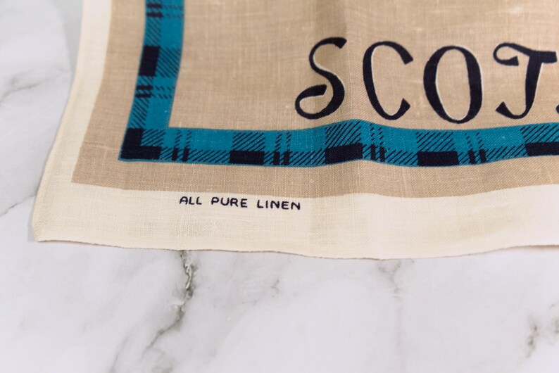 Puede incluir: Un pa&ntilde;o de cocina de lino color canela con un borde de cuadros azul oscuro y azul verdoso. El texto SCOT est&aacute; impreso en letras negras grandes y las palabras ALL PURE LINEN est&aacute;n impresas en letras negras peque&ntilde;as cerca del borde inferior.