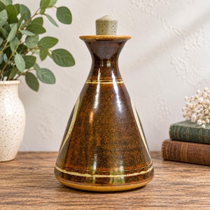 May include: A conical ceramic oil cruet with a glossy dark brown glaze and a speckled light grey stopper. The vessel features a wide base that tapers to a narrow neck, accented by horizontal cream-coloured bands encircling the body.