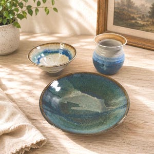 May include: A collection of three ceramic pieces including a small bowl with a blue and white speckled glaze, a tall cylindrical vase with a blue and white gradient finish, and a wide, shallow bowl featuring a deep green and blue reactive glaze.