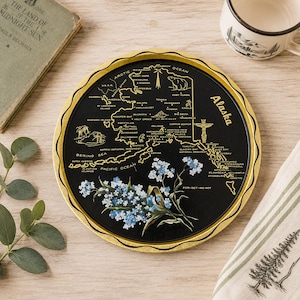 May include: A round black tray with a gold scalloped rim featuring a gold map of Alaska with labeled cities, a polar bear, and a totem pole illustration. A cluster of blue forget-me-not flowers is printed at the bottom with the text Forget-Me-Not.
