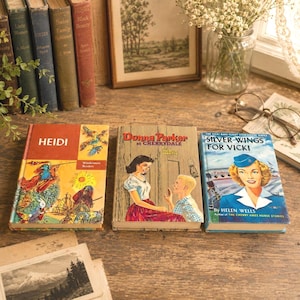 Puede incluir: Tres libros de tapa dura antiguos titulados Heidi, Donna Parker at Cherrydale y Silver Wings for Vicki. Los libros presentan portadas ilustradas coloridas con personajes y texto, exhibidos sobre una superficie de madera junto a otros libros.
