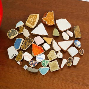 May include: A collection of irregularly shaped, colourful ceramic pieces, resembling sea glass, arranged on a wooden surface. The pieces feature various colours and patterns, including white, blue, orange, and speckled designs. The arrangement suggests a craft or decorative theme.