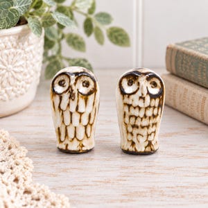 May include: Two ceramic owl figurines featuring a cream-colored glaze with dark brown accents highlighting the carved feather details and circular eyes. These small, upright decorative sculptures have a smooth, glossy finish and a rounded shape.