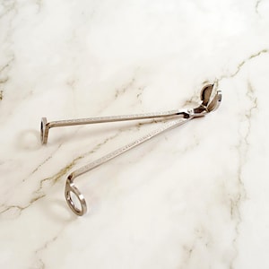May include: Silver candle wick trimmer with a curved blade and long handles. The handles have a decorative etched design and the text "YANKEE CANDLE" is visible. The trimmer is designed to trim candle wicks for a cleaner burn.