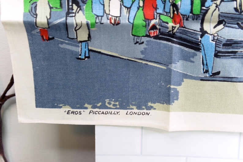Puede incluir: Un pa&ntilde;o de cocina de tela blanca con una colorida ilustraci&oacute;n gr&aacute;fica de personas en una plaza de la ciudad. La escena incluye figuras con abrigos y el texto "EROS" PICCADILLY, LONDON. impreso en negro en la esquina inferior izquierda.