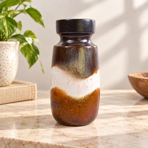 May include: A cylindrical ceramic vase featuring a dark brown neck that transitions into a wide, wavy white band across the centre, followed by a speckled, deep amber-brown base. The surface has a glossy, multi-toned glaze finish.