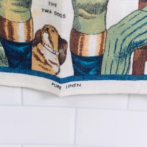 Puede incluir: Un pa&ntilde;o de cocina de lino color crema con una ilustraci&oacute;n tejida de dos perros y el texto THE TWA DOGS. El borde inferior incluye el texto impreso PURE LINEN. El dise&ntilde;o incorpora tonos verdes, marrones y azules sobre una tela texturizada.
