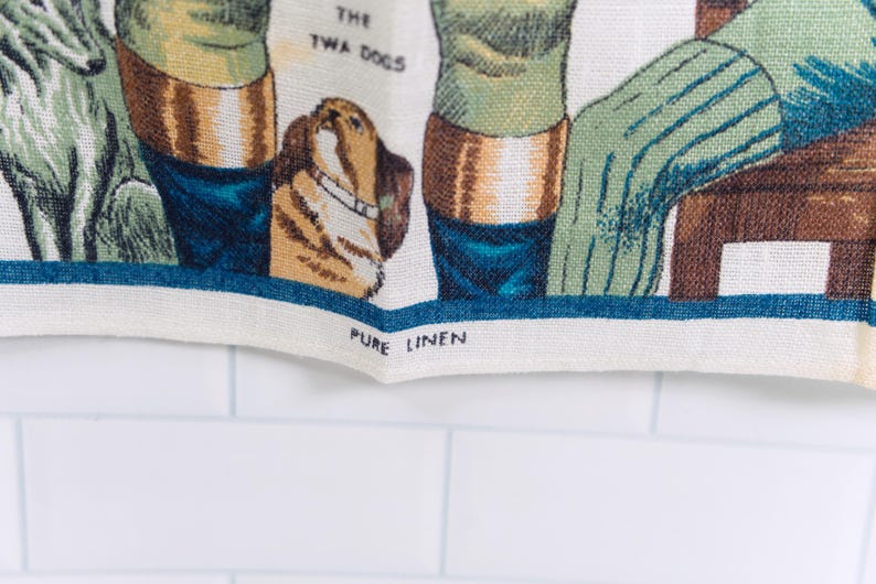 Puede incluir: Un pa&ntilde;o de cocina de lino color crema con una ilustraci&oacute;n tejida de dos perros y el texto THE TWA DOGS. El borde inferior incluye el texto impreso PURE LINEN. El dise&ntilde;o incorpora tonos verdes, marrones y azules sobre una tela texturizada.