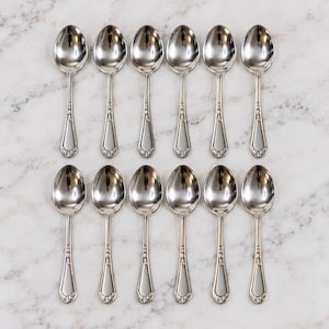 May include: A set of twelve polished silver-tone metal spoons arranged in two rows of six. Each spoon features a smooth oval bowl and a slender handle with a decorative embossed floral pattern at the base of the stem and the tip of the handle.