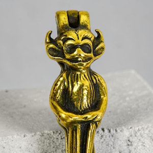 May include: A brass figurine of a goblin-like creature with a smiling face, large ears, and a crown. The figurine has a textured surface and is standing upright. The goblin is holding its hands together in front of its body.