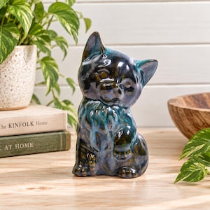 Peut inclure: Une figurine de chat en céramique avec un vernis brillant aux tons multiples, mêlant des nuances de bleu profond, de sarcelle et de brun foncé. Le chat est représenté assis avec une patte avant légèrement levée, offrant une finition lisse.