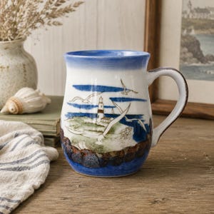 Peut inclure: Une tasse en céramique avec une base blanche et un bord bleu, ornée d'une scène côtière peinte à la main représentant un phare, un rivage rocheux et des mouettes en vol. Le motif entoure le corps de la tasse, qui a une forme incurvée et une anse.