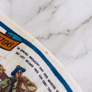 Puede incluir: Un pa&ntilde;o de cocina de tela blanca con un borde azul y una ilustraci&oacute;n impresa de una persona con sombrero azul. El texto dice BURNS - 1796 y AE SPRING BROUGHT OFF HER MASTER M BUT LEFT BEHIND HER AIN GREY TAIL. La tela muestra manchas de color marr&oacute;n claro.