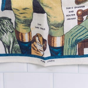 Puede incluir: Un pa&ntilde;o de cocina de lino blanco con una colorida ilustraci&oacute;n impresa de figuras y animales. El texto incluye 'THE TWA DOGS', 'TAM O SHANTER AND SOUTER JOHNNY' y 'PURE LINEN'. El dise&ntilde;o muestra un perro gris, un perro marr&oacute;n y piernas.