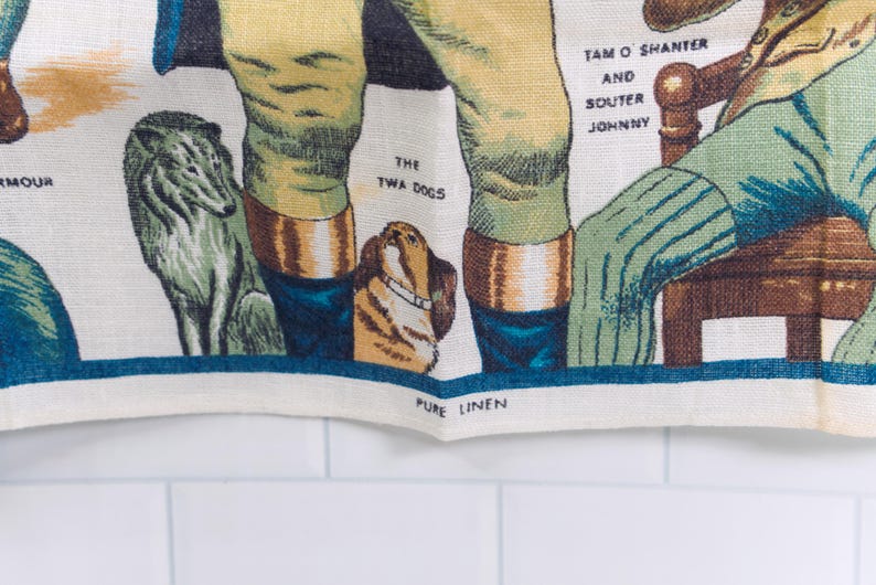 Puede incluir: Un pa&ntilde;o de cocina de lino blanco con una colorida ilustraci&oacute;n impresa de figuras y animales. El texto incluye 'THE TWA DOGS', 'TAM O SHANTER AND SOUTER JOHNNY' y 'PURE LINEN'. El dise&ntilde;o muestra un perro gris, un perro marr&oacute;n y piernas.
