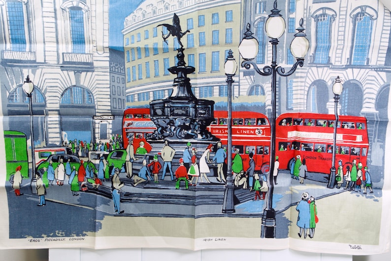 Puede incluir: Un pa&ntilde;o de cocina de recuerdo con una ilustraci&oacute;n colorida de la estatua de Eros en Piccadilly Circus, Londres. La escena incluye autobuses rojos de dos pisos, farolas y peatones. El texto inferior dice Eros Piccadilly London.