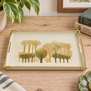 May include: A rectangular serving tray with a cream-coloured base and a gold-toned metal frame at the corners. The centre features a graphic illustration of a forest with trees in shades of olive green and metallic gold, set against thin horizontal lines.