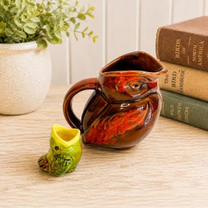 May include: A glossy brown ceramic owl-shaped creamer pitcher with orange accents and a handle, paired with a smaller, bright green ceramic bird-shaped creamer with a yellow interior, both featuring detailed molded feathers and avian facial features.