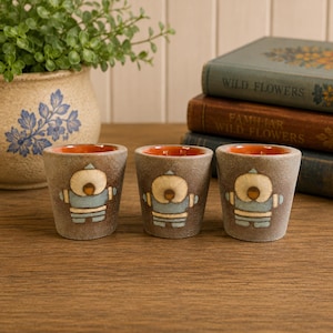 May include: Three small, textured, grey ceramic cups with a matte finish. Each cup features a central embossed graphic of a figure wearing a hooded parka in shades of light blue and cream. The interior of each cup is glazed in a vibrant orange.
