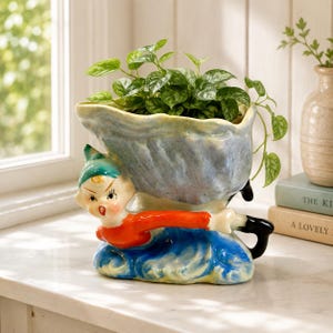 May include: A ceramic planter shaped like a whimsical elf figure pushing a wheelbarrow, featuring a glossy finish in shades of blue, orange, and black. The elf has a painted face and green hat, while the wheelbarrow holds a green leafy plant.