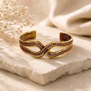 Peut inclure: Bracelet manchette doré au design complexe. Le bracelet présente des bandes entrelacées avec une texture en forme de chaîne et un accent brun-rougeâtre chaud. Le design ouvert permet de le porter facilement. Un bijou élégant.