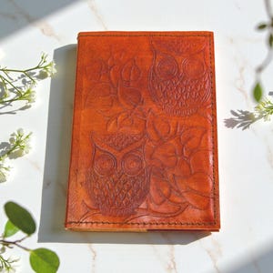 May include: A rectangular, orange-brown leather journal with embossed owl and floral designs. The cover features two owls and leaves, with a stitched border. The journal is closed and sits on a white surface with sunlight.