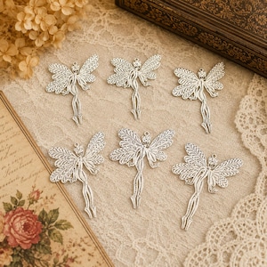 May include: Six silver fairy charms with intricate openwork wings and slender bodies. Each fairy has a small loop for hanging. The charms are displayed on a neutral-toned fabric.