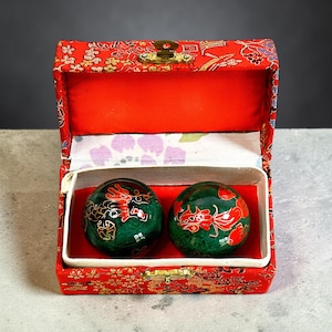 May include: A red, patterned box with a gold clasp, open to reveal two green, patterned spheres. The spheres feature red dragon designs. The box's interior is red with a white and purple floral patterned lining.