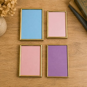 May include: Four rectangular picture frames with thin gold-toned metal borders. Each frame contains a solid-coloured insert: one light blue, two light pink, and one purple. The frames are arranged in a grid pattern on a flat wooden surface.