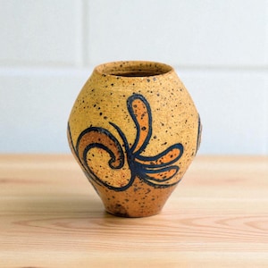 May include: A small, speckled ceramic vase with a rounded body and a narrow opening. The vase is primarily tan with dark brown and orange abstract floral designs. The vase sits on a wooden surface.