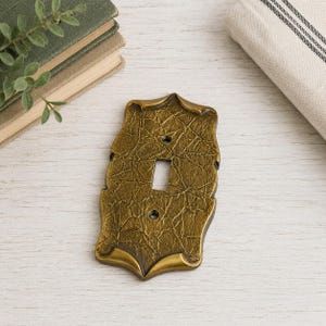 May include: A rectangular light switch wall plate with a textured, antique brass finish. The plate features a central vertical toggle opening and two screw holes, one above and one below the opening, with decorative scalloped edges.