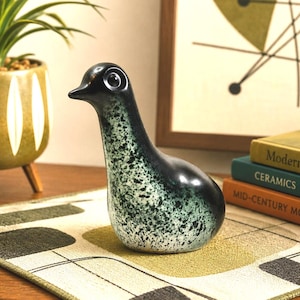 May include: A ceramic duck figurine with a speckled green and black finish. The duck has a long neck and a rounded body. The figurine is displayed on a patterned surface, with a plant and books in the background.