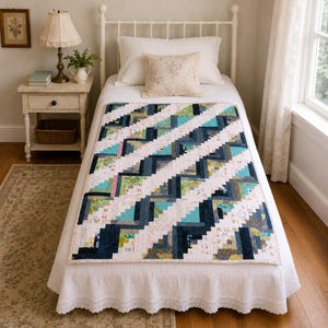 May include: A rectangular quilt featuring a geometric log cabin pattern with alternating diagonal stripes of dark navy blue, teal, and light floral patterned fabric. The quilt is spread flat across a white bed frame with a white bed skirt.