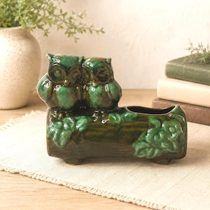Peut inclure: Une jardinière en céramique en forme de bûche avec deux hiboux perchés sur le dessus. La pièce présente un vernis brillant vert foncé et marron avec des détails de feuilles moulées sur les côtés. Les hiboux ont de grands yeux ronds.