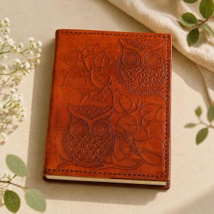 May include: A rectangular notebook with a tan leather cover featuring an embossed design of two owls perched among leafy branches. The cover has visible stitching along the edges and a smooth, polished finish with a warm, medium-brown tone.