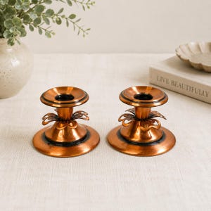 Peut inclure: Deux bougeoirs en métal de couleur cuivre assortis, dotés d'une large base circulaire, d'une tige centrale avec des accents décoratifs en forme de pétales et d'un bord supérieur évasé. La finition présente un éclat métallique poli.