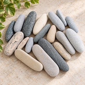 May include: A collection of smooth, elongated river stones in various shades of gray, charcoal, beige, and cream. The stones are oval-shaped and vary in size, featuring natural textures and speckled patterns across their surfaces.