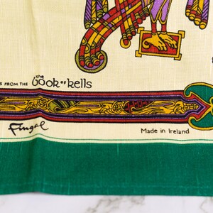 Puede incluir: Un tejido de color crema con ilustraciones coloridas del Libro de Kells, que incluyen intrincados patrones rojos, amarillos y morados. El texto dice Illustrations from the Book of Kells, Fingal y Made in Ireland, con un borde verde.
