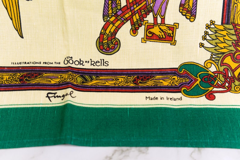 Puede incluir: Un tejido de color crema con ilustraciones coloridas del Libro de Kells, que incluyen intrincados patrones rojos, amarillos y morados. El texto dice Illustrations from the Book of Kells, Fingal y Made in Ireland, con un borde verde.