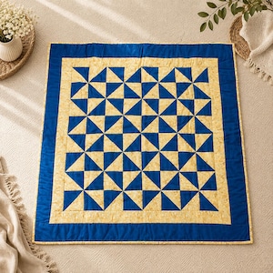 May include: A square quilt featuring a repeating geometric pattern of blue and cream-coloured triangles arranged in a grid. The quilt is framed by a wide solid blue border and a thinner inner border of cream fabric with a subtle mottled texture.