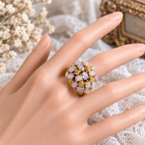 May include: A gold ring featuring a cluster of multiple small, oval-shaped opals with iridescent flashes of color. The setting is composed of intricate, textured gold metal elements that hold the stones in a rounded, dome-like arrangement.