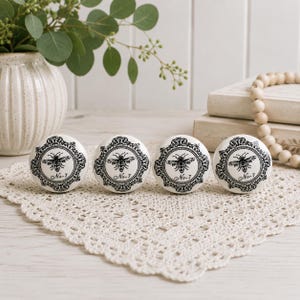 May include: Four round white ceramic knobs featuring a black printed design of a bee centred within an ornate circular frame. Each knob includes the text No-7 printed in a script font below the bee illustration, creating a uniform set.